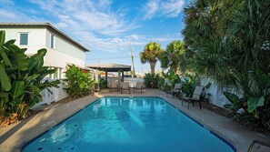A swimming pool with a covered seating area, surrounded by lush greenery and a white building. - Chasing Sunshine by Avantstay Shared Pool + Gazebo + Covered Patio Walk to the Beach (Port Aransas)