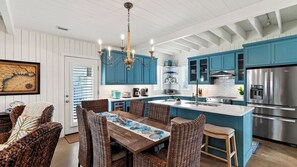 A modern kitchen with blue cabinets, stainless steel appliances, and a dining area with wicker chairs and a wooden table. - Chasing Sunshine by Avantstay Shared Pool + Gazebo + Covered Patio Walk to the Beach (Port Aransas)