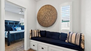 Una habitación con un sofá azul oscuro, un adorno circular tejido en la pared, una puerta blanca y una ventana con persianas.
