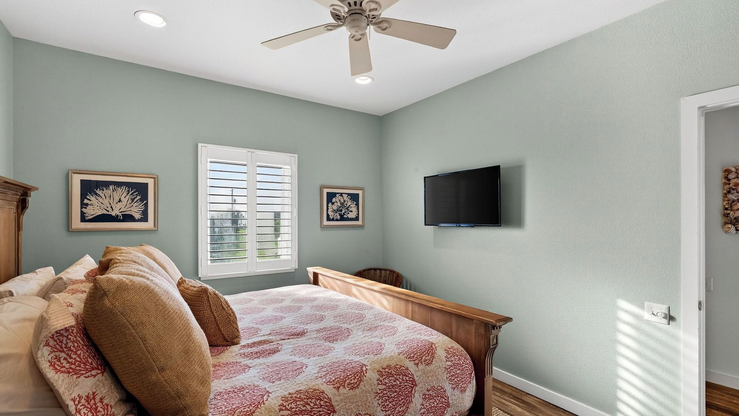 Une chambre à coucher avec un lit, un ventilateur de plafond, une fenêtre avec des stores et un téléviseur fixé au mur.