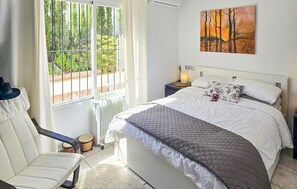 A bedroom with a bed, a chair, a nightstand, a radiator, and a window with a view of greenery. - Cozy home in Viñuela with private swimming pool, can be inside or outside (Viñuela)