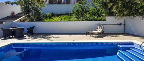 A swimming pool with steps leading into it, surrounded by outdoor furniture and a house in the background.