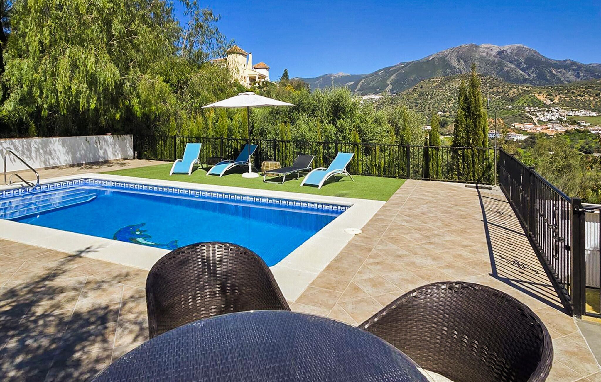 Una piscina con sillas de descanso y una mesa, rodeada por una valla y árboles, con vistas a la montaña al fondo.