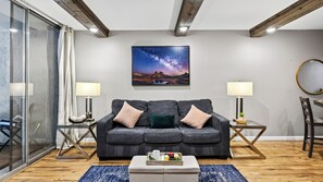 Ein modernes Wohnzimmer mit einem dunkelgrauen Sofa, zwei Beistelltischen mit Lampen, einem Couchtisch mit einer Snackplatte und einem großen gerahmten Bild des Nachthimmels an der Wand.