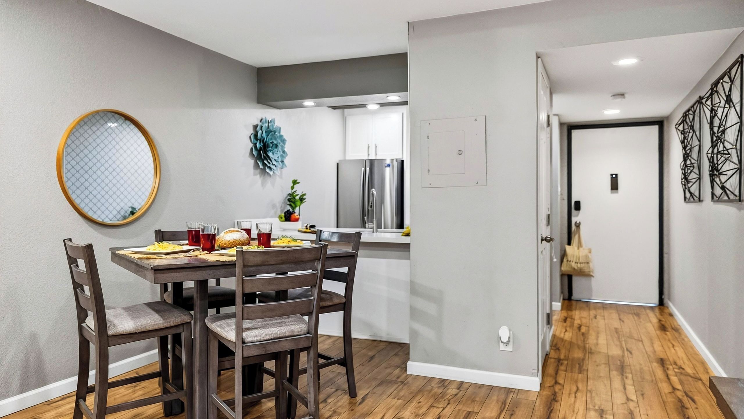 Comedor con mesa dispuesta para cuatro personas, sillas, un espejo redondo y un adorno en la pared.