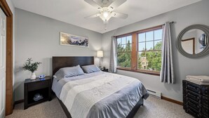 Un dormitorio con cama, mesita de noche, lámpara, ventana con vistas, ventilador de techo y espejo.