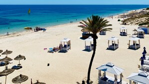 A beach with clear blue water, sun loungers, and beach huts. - Résidence Nesrine Cozy Stay in Yasmine  (Port El Kantaoui)