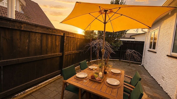 A wooden patio table with a yellow umbrella, surrounded by green chairs, set against a backdrop of a wooden fence and a white building. - Maison Millie - Central, Pet-Friendly, Sleeps 4 (Rotorua)