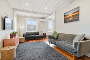 Ein modernes Wohnzimmer mit einem grauen Sofa, einem schwarzen Ledersofa und einem grauen Sessel. An der Wand hängt ein Gemälde, auf der Anrichte steht eine Pflanze und auf dem Boden steht ein gewebter Korb.