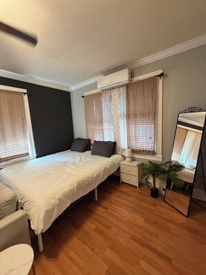 Ein Schlafzimmer mit Bett, Nachttischen, einem Spiegel, einer Topfpflanze und einem Fenster mit Vorhängen.