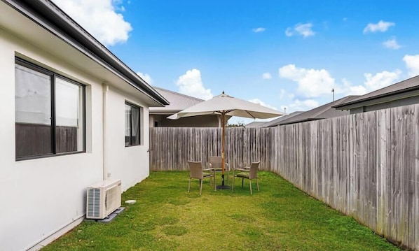 A backyard with a wooden fence, a white house, a table with chairs, and an umbrella. - Stylish 4BR Sleeps Up To 8 Near Theme Parks (Coomera)