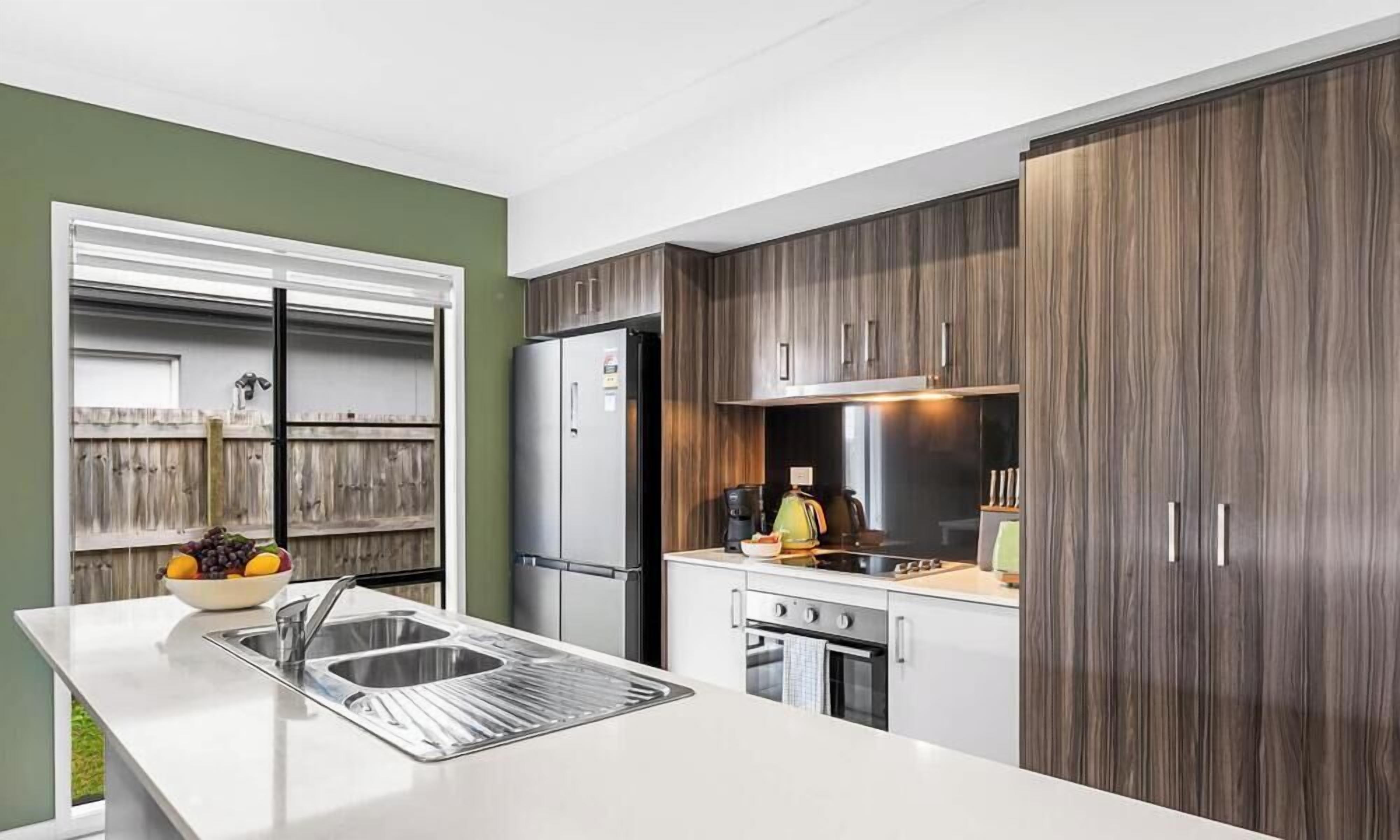 A modern kitchen with a stainless steel refrigerator, a built-in oven, and a sink with a bowl of fruit.
