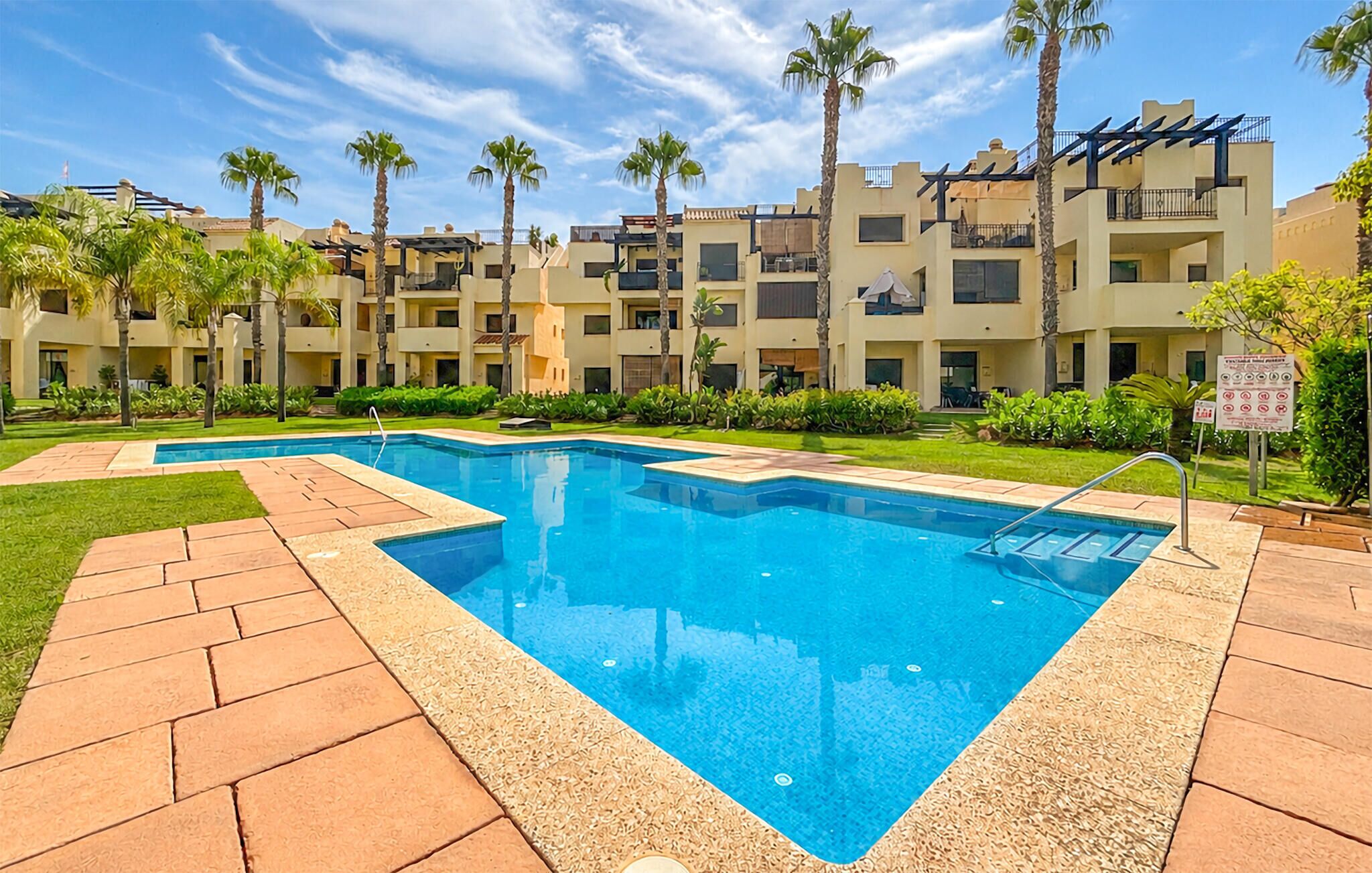 Una piscina con plataforma de cerámica, rodeada de palmeras y un edificio de varios pisos con balcones.