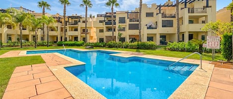Una piscina con plataforma de cerámica, rodeada de palmeras y un edificio de varios pisos con balcones.