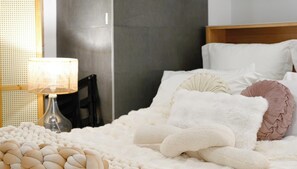 Uma cama bem arrumada com almofadas brancas e bege, um cobertor branco fofo e um abajur em uma mesinha de cabeceira.