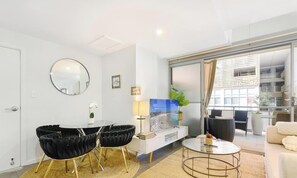 A modern living room with a flat-screen TV, a round mirror, and a dining area with a white table and black chairs. - KozyGuru | Parramatta | Prime City Centre Living (Parramatta)