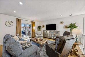 A modern living room with a sofa, armchair, and a coffee table. There is a television mounted on the wall and a window with curtains. - Remodeled & modern coastal escape duplex with pool, close to beach (Fernandina Beach)