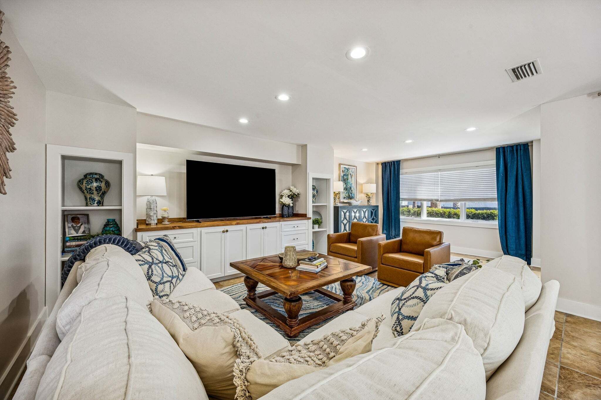 A living room with a sofa, coffee table, and two chairs. There is a flat-screen TV mounted on the wall, a window with curtains, and a ceiling vent.