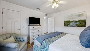 Un dormitorio con cama, televisor en una cómoda, una silla y un cuadro en la pared.