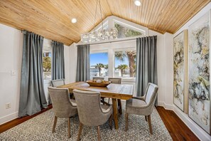 A dining area with a wooden table, four upholstered chairs, a chandelier, and large windows with curtains. - Spectacular renovated duplex with pool near beach (Fernandina Beach)