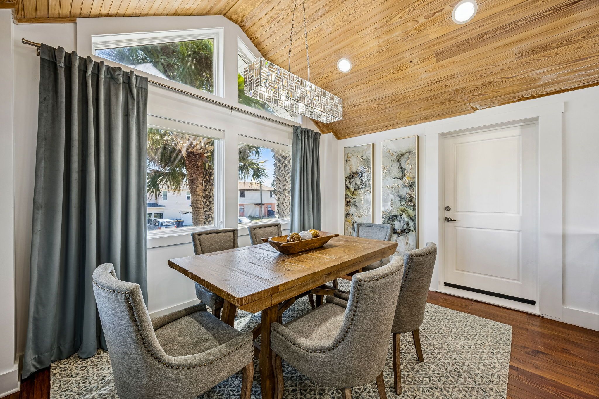 A dining area with a wooden table and four upholstered chairs, a window with curtains, and a door with artwork on the wall.