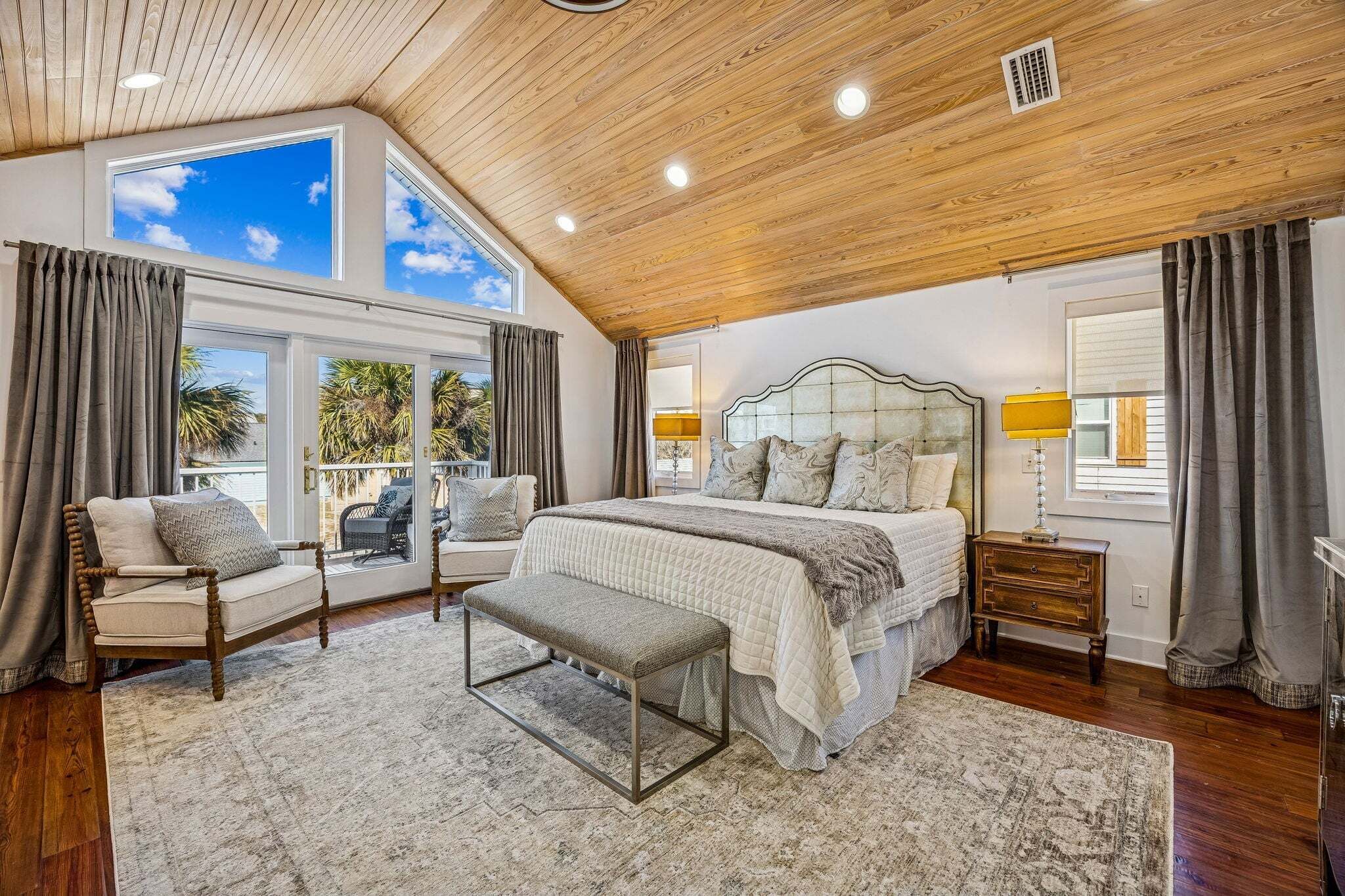 A bedroom with a large bed, a bench, a chair, a nightstand, and a window with curtains.