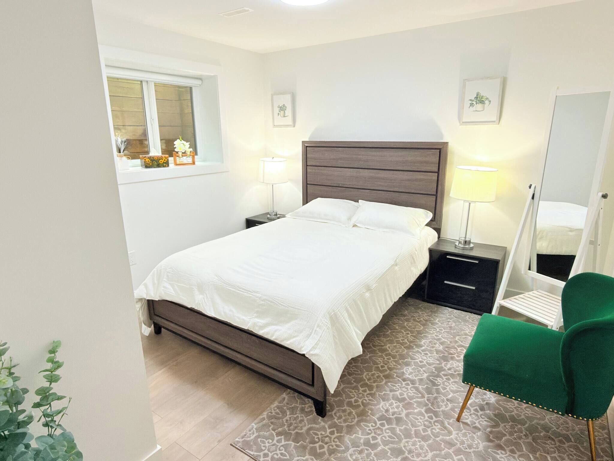 Une chambre à coucher avec un lit, des tables de chevet, un miroir, un fauteuil vert et une fenêtre donnant sur un beau paysage.
