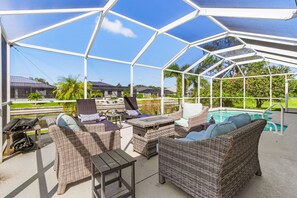 A covered outdoor area with wicker furniture, a pool, and a view of houses and greenery. - Sunset Villa 1035 Modern Waterfront Heated Pool Home w/ Fire Pit (Cape Coral)