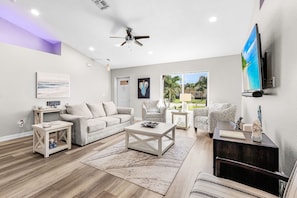 A modern living room with a sofa, coffee table, and patterned chairs. A ceiling fan and a flat-screen TV are mounted on the wall. - Sunset Villa 1035 Modern Waterfront Heated Pool Home w/ Fire Pit (Cape Coral)