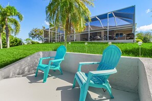 A modern house with a glass facade, outdoor seating area with turquoise chairs, and a well-maintained lawn. - Sunset Villa 1035 Modern Waterfront Heated Pool Home w/ Fire Pit (Cape Coral)