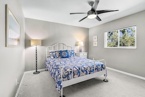 A bedroom with a bed, two bedside tables, a ceiling fan, and a window with a view of trees. - Sunset Villa 1035 Modern Waterfront Heated Pool Home w/ Fire Pit (Cape Coral)