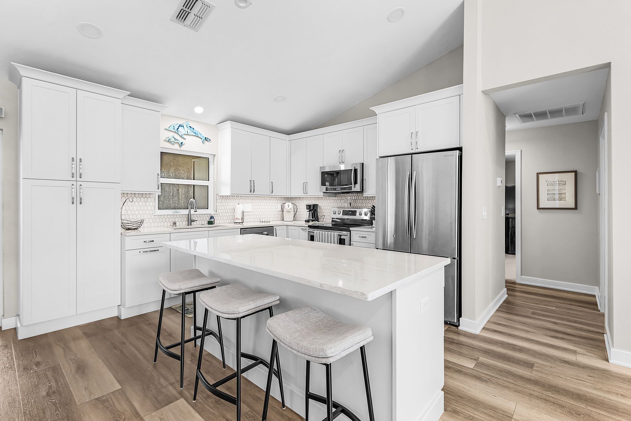 A modern kitchen with white cabinetry, a central island with bar stools, stainless steel appliances, and a tiled backsplash.