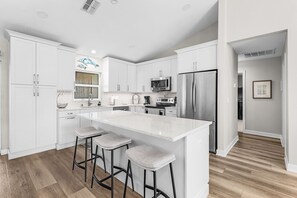 A modern kitchen with white cabinetry, a central island with bar stools, stainless steel appliances, and a tiled backsplash. - Sunset Villa 1035 Modern Waterfront Heated Pool Home w/ Fire Pit (Cape Coral)