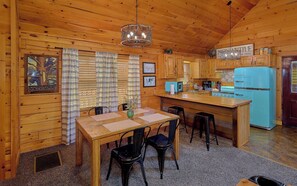 Ein Holzblockbau-Innenraum mit Esstisch, Stühlen, einer Küchenzeile mit blauem Kühlschrank und einer Hängeleuchte.