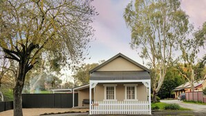 A single-story house with a front porch, a fenced yard, and a tree in the foreground. - The Butcher's Cottage | New Barossa Valley Stay (Williamstown)