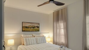 A hotel room with a bed, two bedside lamps, a ceiling fan, a painting on the wall, and a window with curtains. - The Butcher's Cottage | New Barossa Valley Stay (Williamstown)