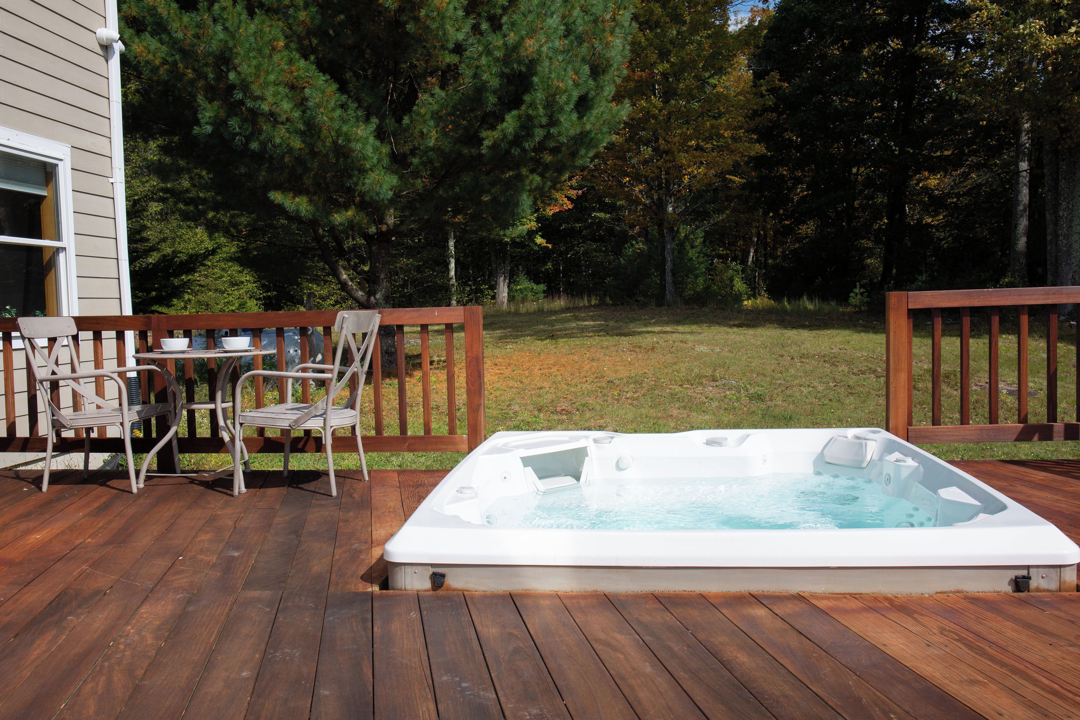 Un bain à remous sur une terrasse en bois, agrémentée de chaises et d’une table, le tout entouré d’une clôture en bois et d’arbres.
