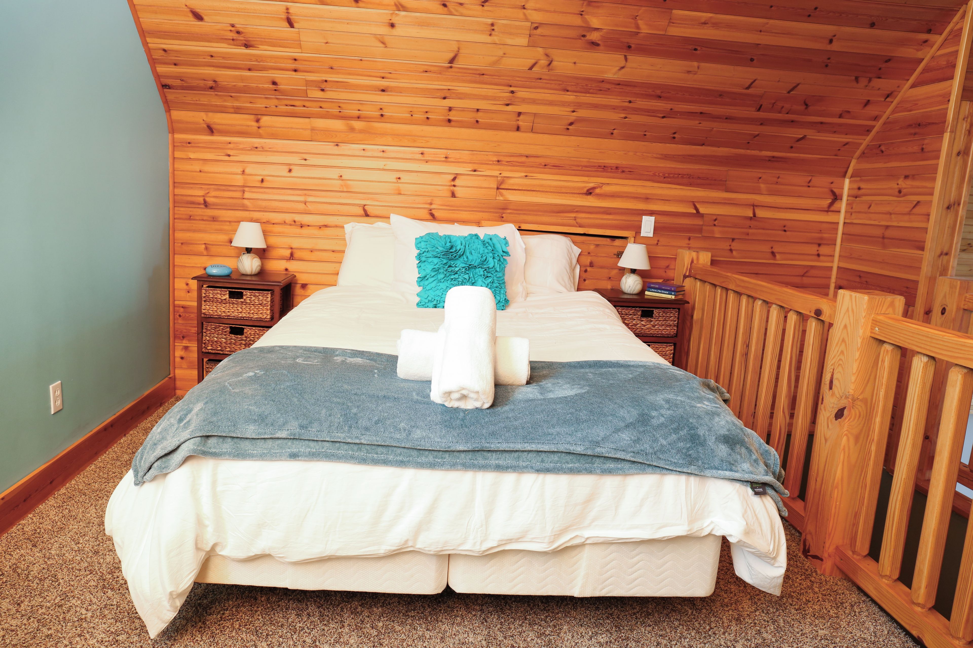Une chambre avec un plafond en pente, des murs en bois, un lit recouvert d’une couverture bleue, deux tables de chevet avec des lampes et une rambarde en bois.