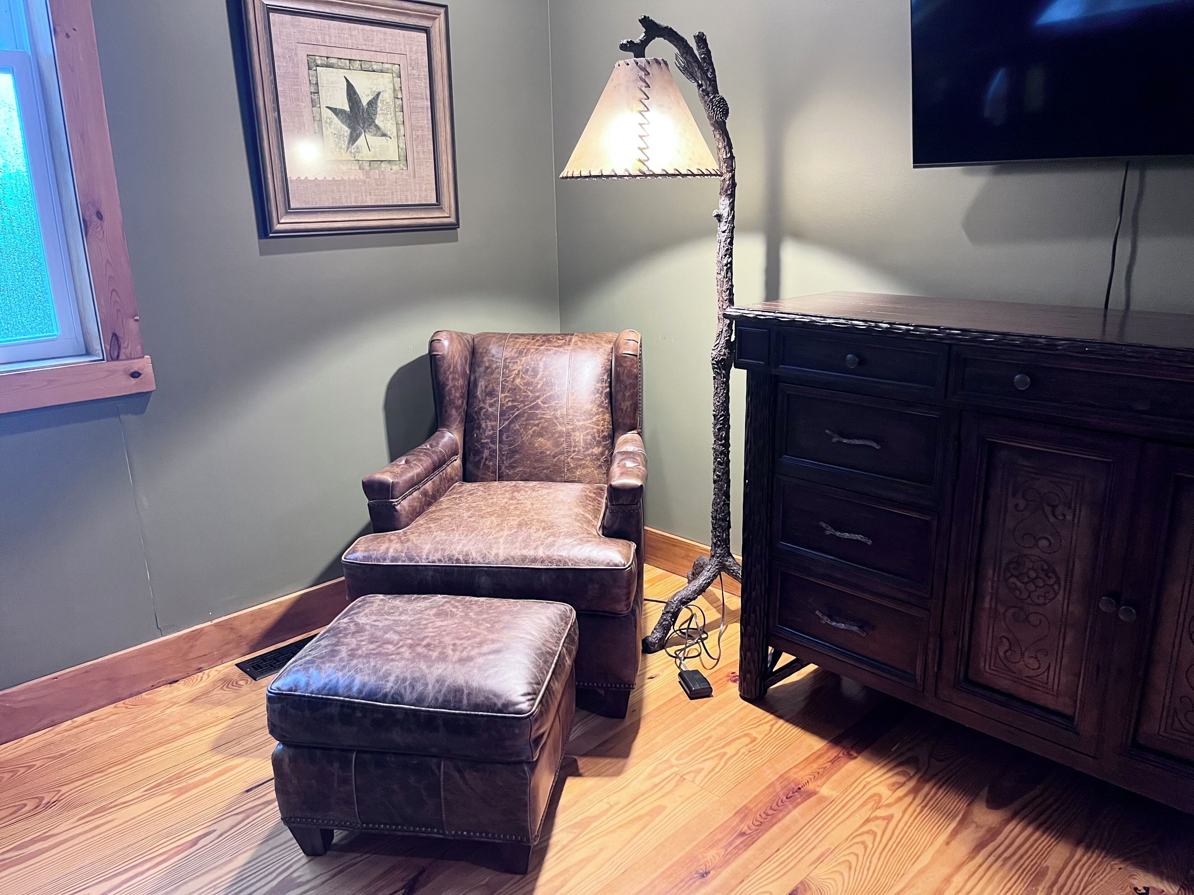 Une pièce comprenant un fauteuil en cuir, un pouf assorti, une armoire en bois, une œuvre d’art encadrée et un lampadaire.