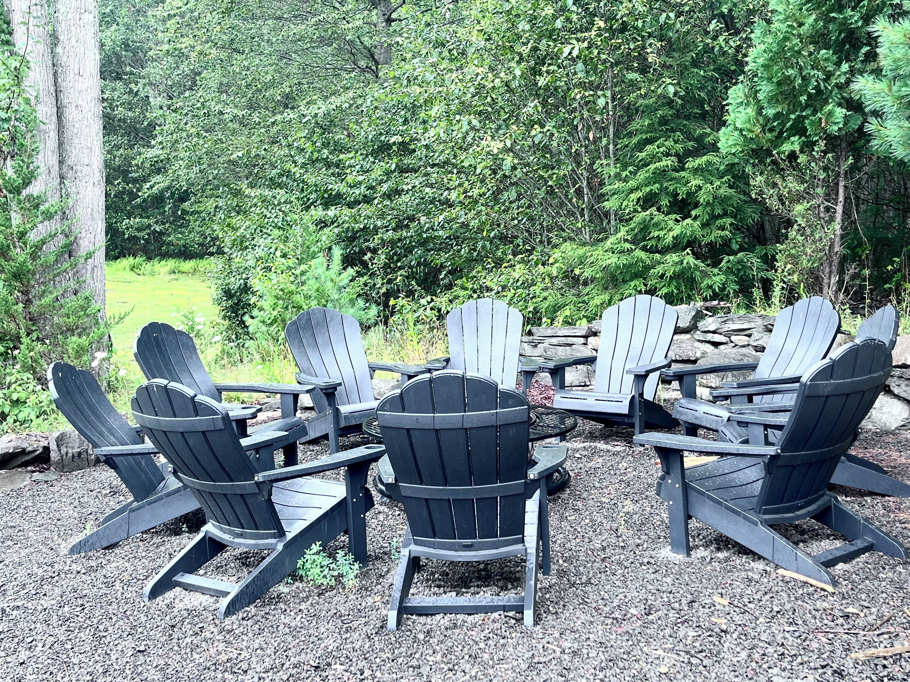 Un groupe de chaises Adirondack disposées autour d’une table ronde dans un espace boisé, le sol étant recouvert de gravier.