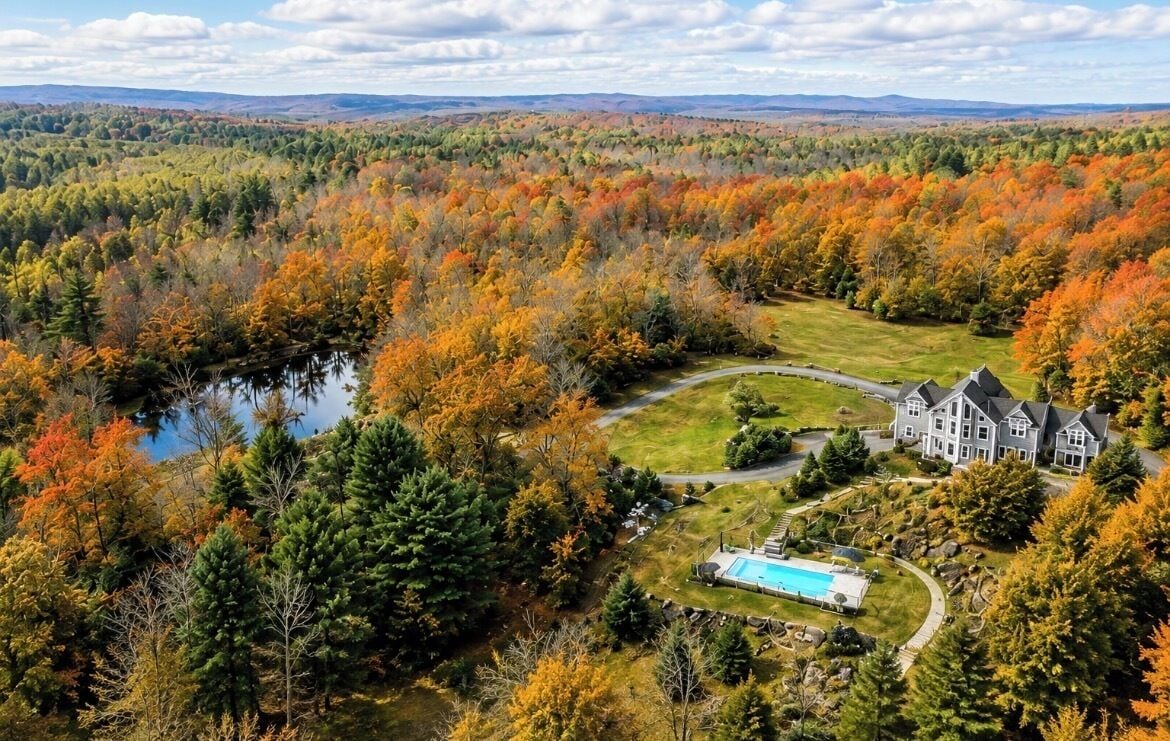 Une grande maison avec une piscine, entourée d’une zone boisée aux couleurs automnales.