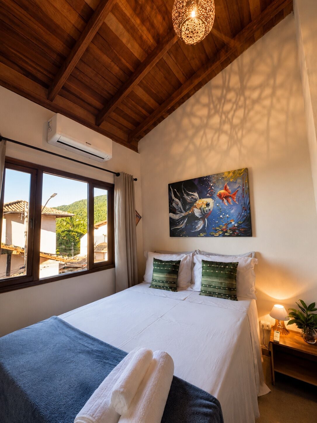 A bedroom with a wooden ceiling, a bed with white bedding, green pillows, a painting of fish, and a window with a view of buildings and greenery.
