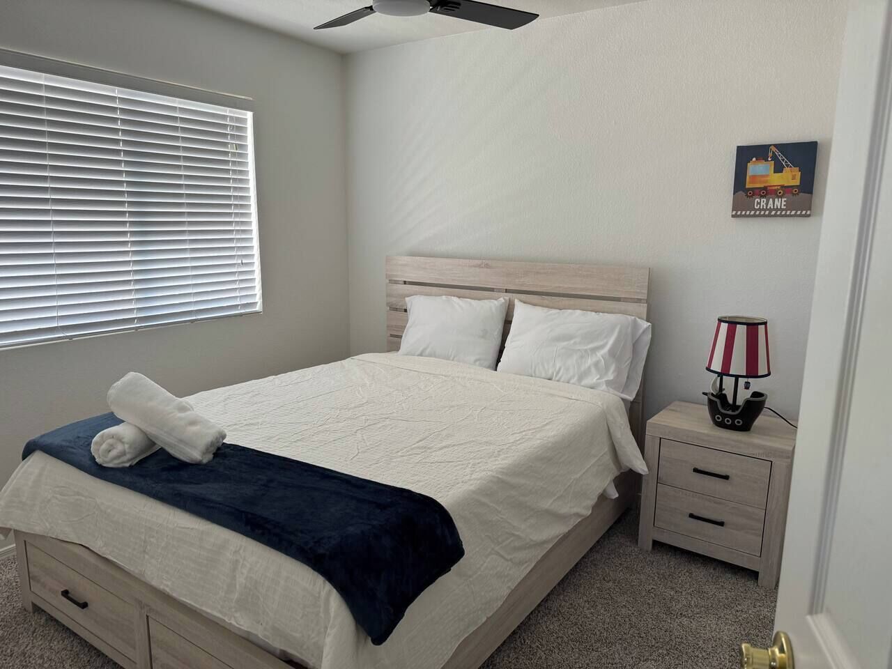 A bedroom with a bed, a nightstand, a lamp, a window with blinds, and a wall decoration.