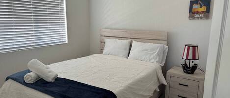 A bedroom with a bed, a nightstand, a lamp, a window with blinds, and a wall decoration.