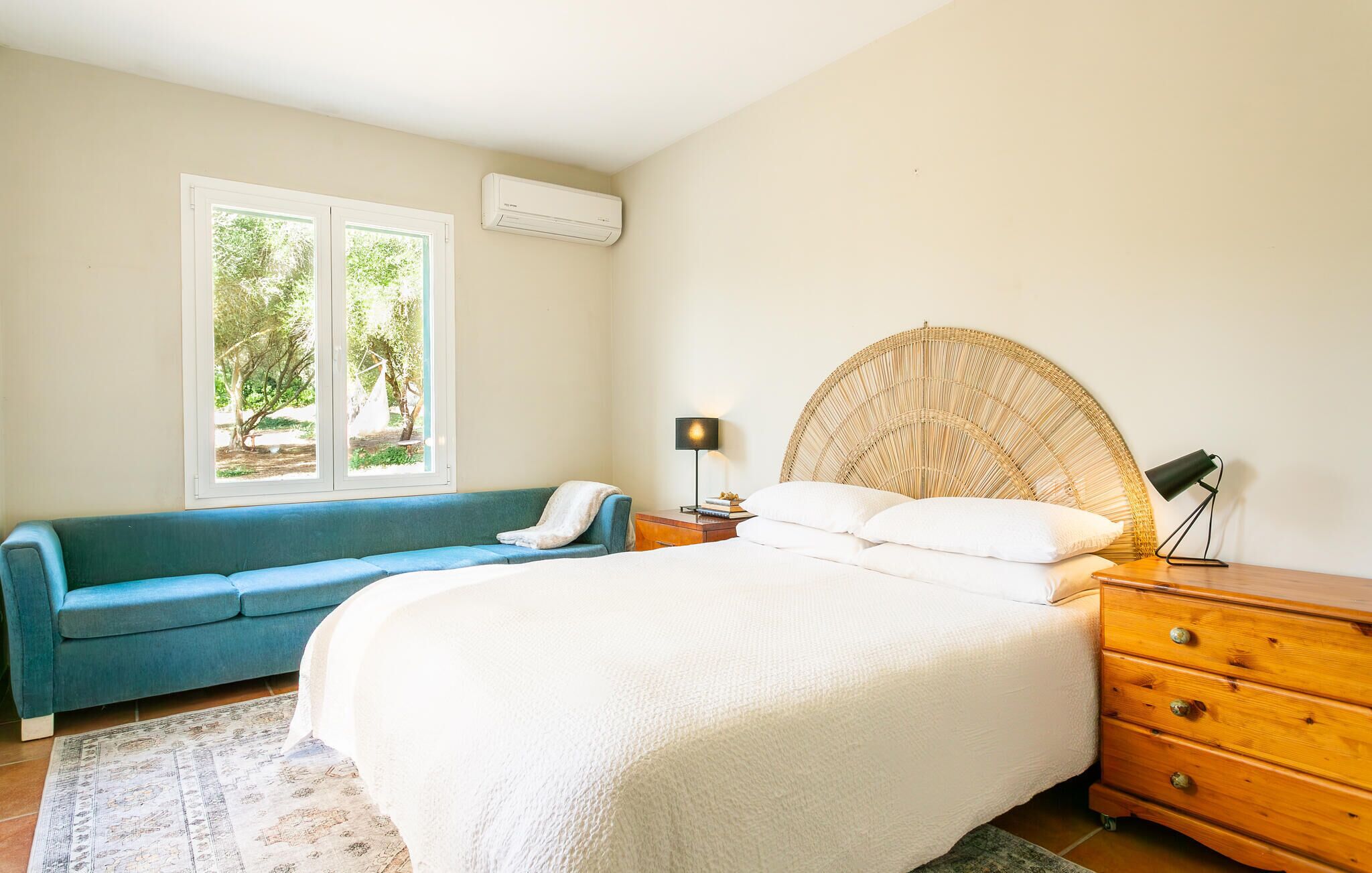 A bedroom with a bed, a blue sofa, a nightstand with a lamp, and a window with a view of greenery.