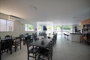 A spacious dining area with multiple tables and chairs, a kitchen area with a counter, and large windows allowing natural light.