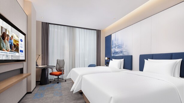 Une chambre d’hôtel avec deux lits, un bureau, une chaise et un écran de télévision diffusant une publicité pour l’hôtel.