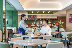 Interior de un restaurante con un camarero atendiendo a un cliente. Las mesas están dispuestas con sillas, y hay elementos decorativos en las paredes.