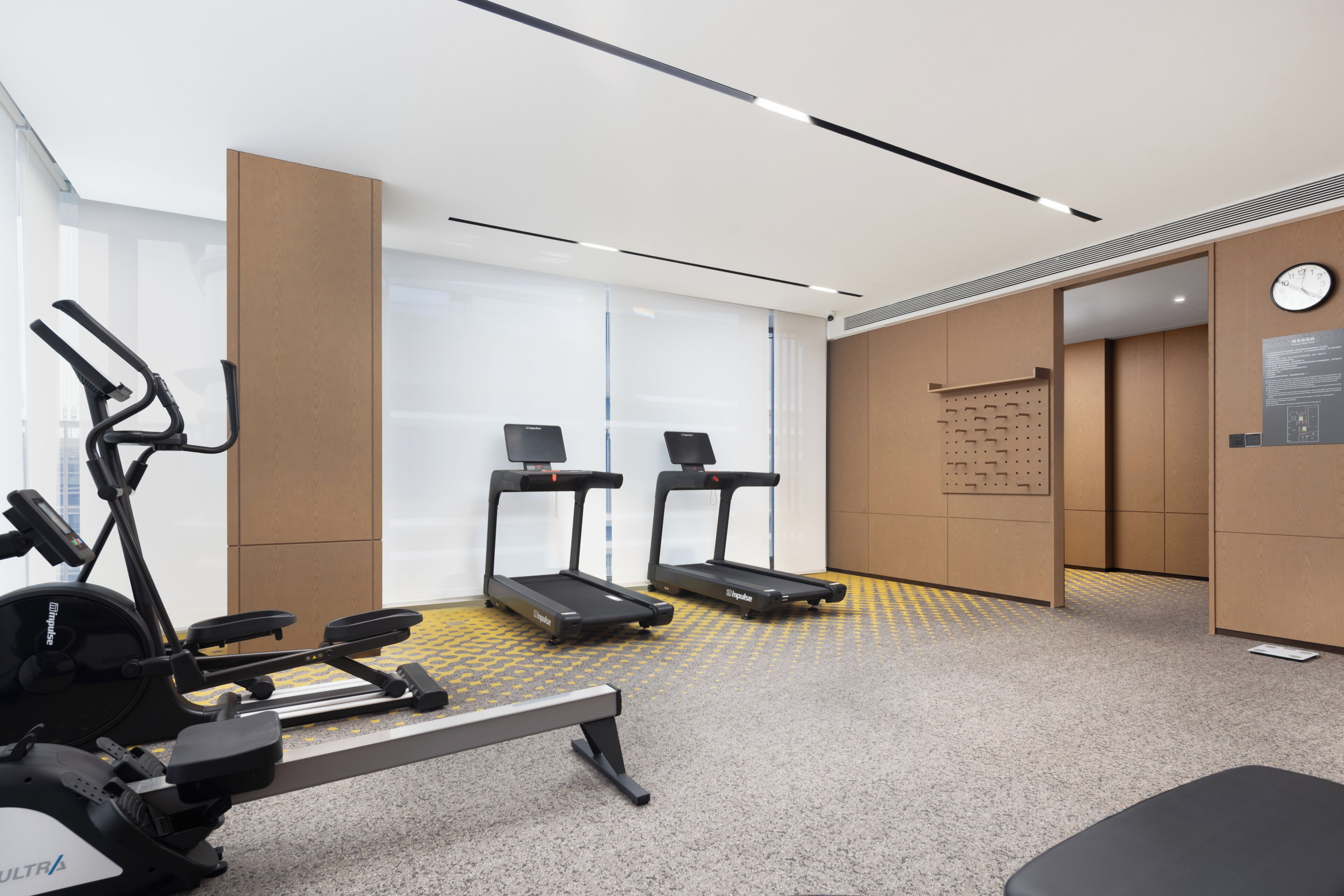 A modern gym with cardio equipment, a wall-mounted clock, and a bulletin board.