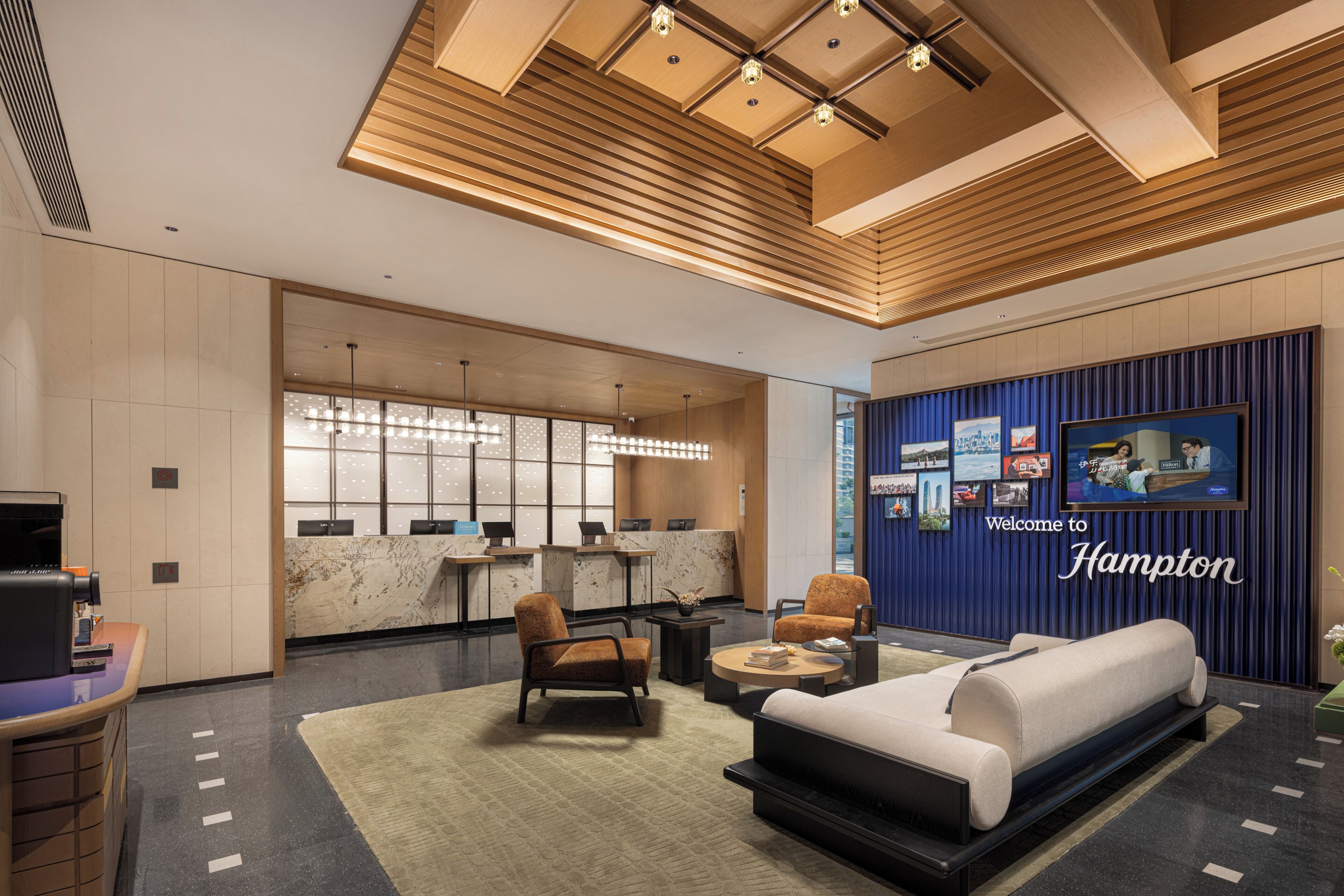 A modern hotel lobby with a reception desk, seating area, and a large screen displaying the hotel's name.